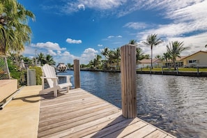A wooden dock with white chairs, a calm water body, and palm trees in the background.