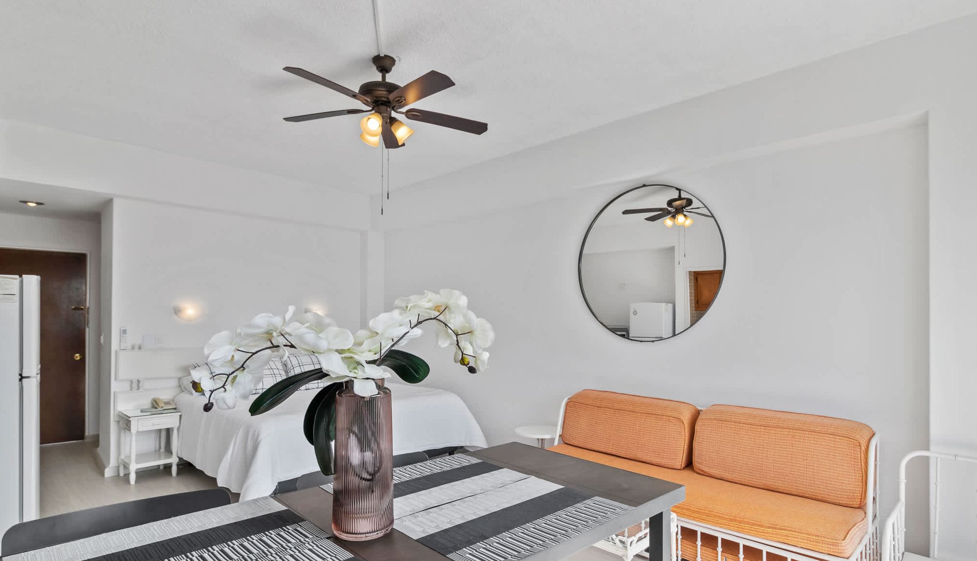 A modern hotel room with a bed, a sofa, a dining table with a vase of flowers, a ceiling fan, and a mirror on the wall.