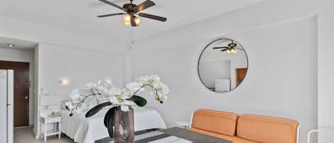 A modern hotel room with a bed, a sofa, a dining table with a vase of flowers, a ceiling fan, and a mirror on the wall.
