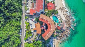 An aerial view of a resort with a circular building, a beach, and a clear blue sea.