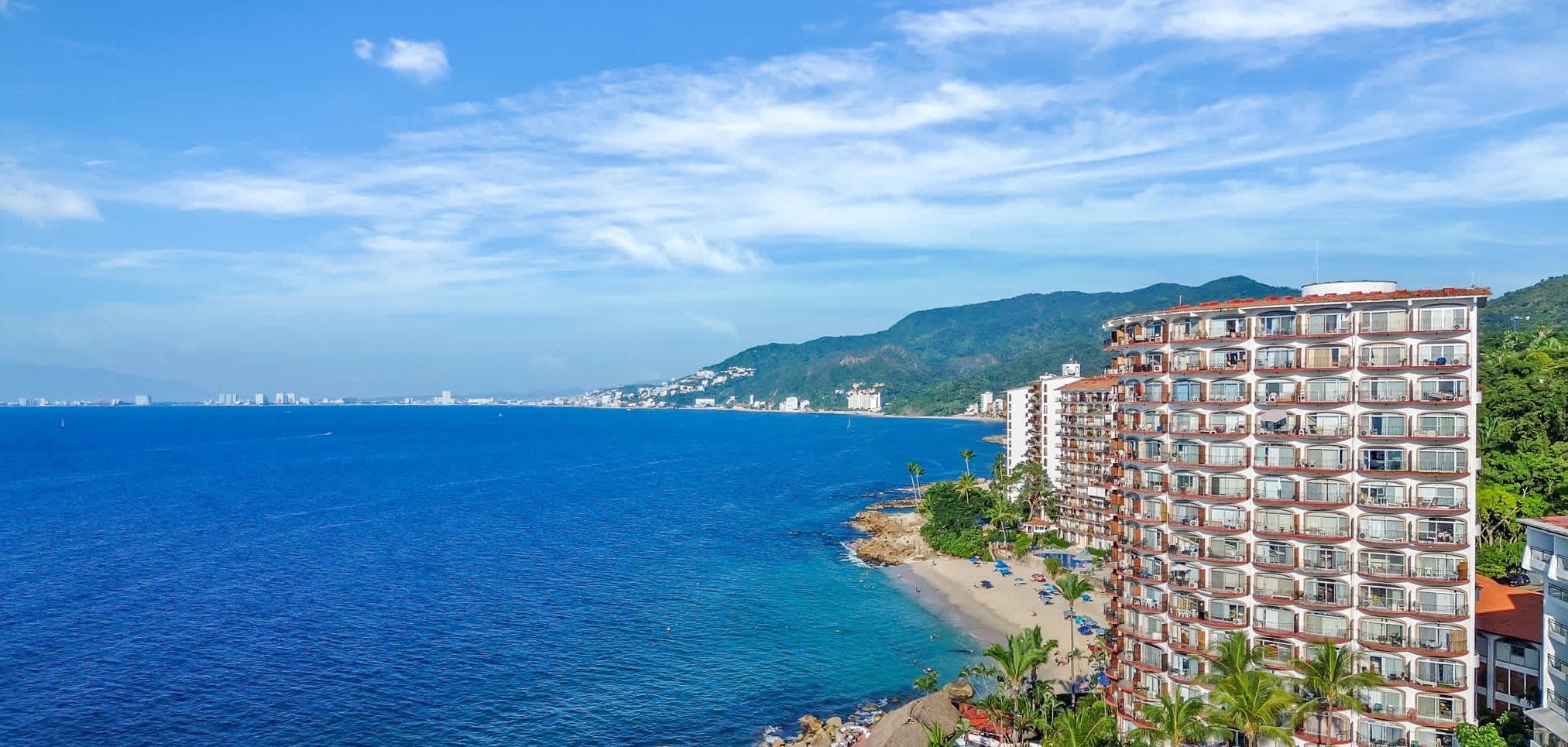 A coastal hotel with multiple balconies, a beachfront, and a view of the sea and distant cityscape.