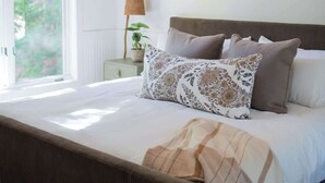 Een slaapkamer met een houten plafond, een bed met veel kussens, een nachtkastje met een lamp, en een raam met uitzicht op groen.