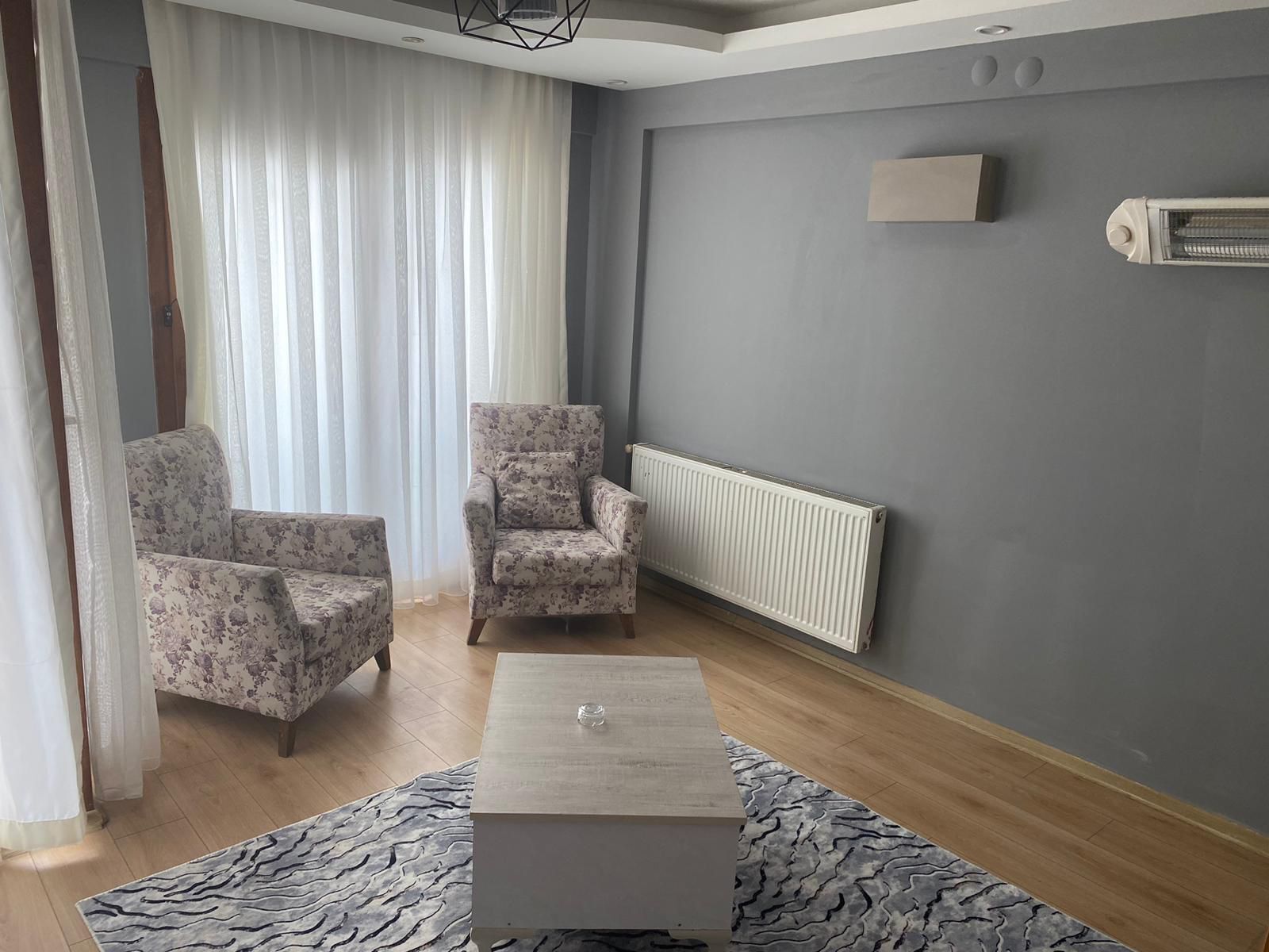 A living room with two floral armchairs, a small coffee table, a radiator, and a window with sheer curtains.