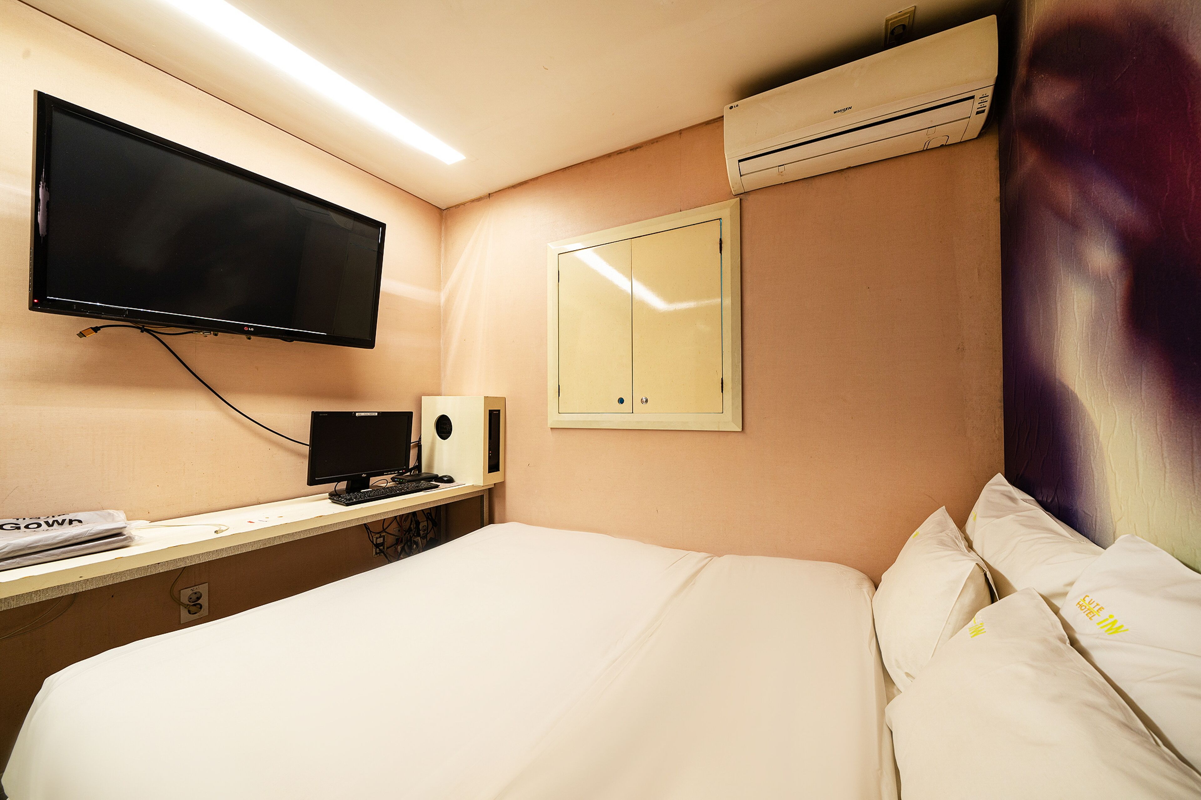 A hotel room with a bed, a flat-screen TV mounted on the wall, a desk with a computer, and an air conditioning unit above the desk.