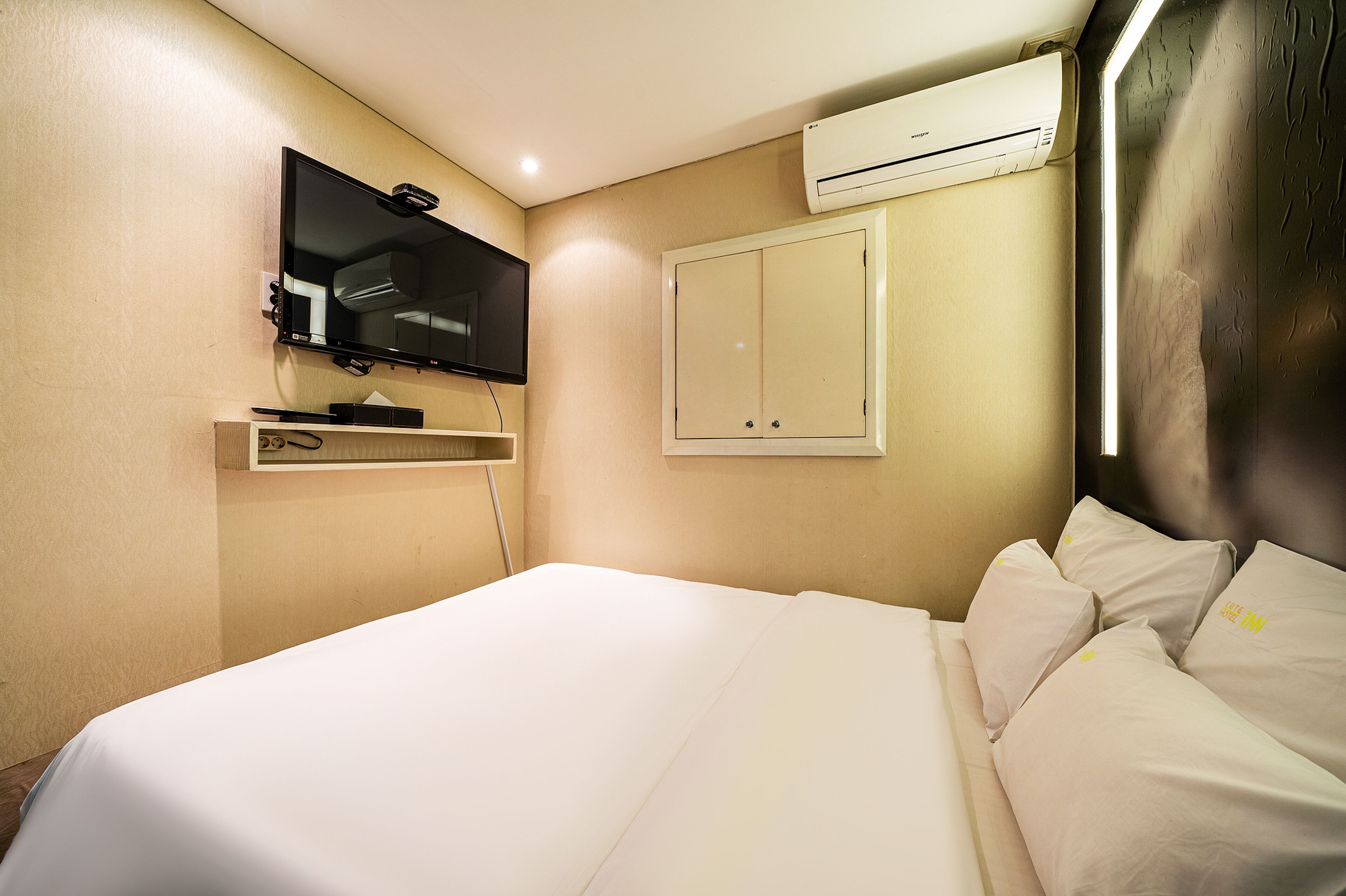 A hotel room with a bed, a mounted television, an air conditioning unit, and a wardrobe.