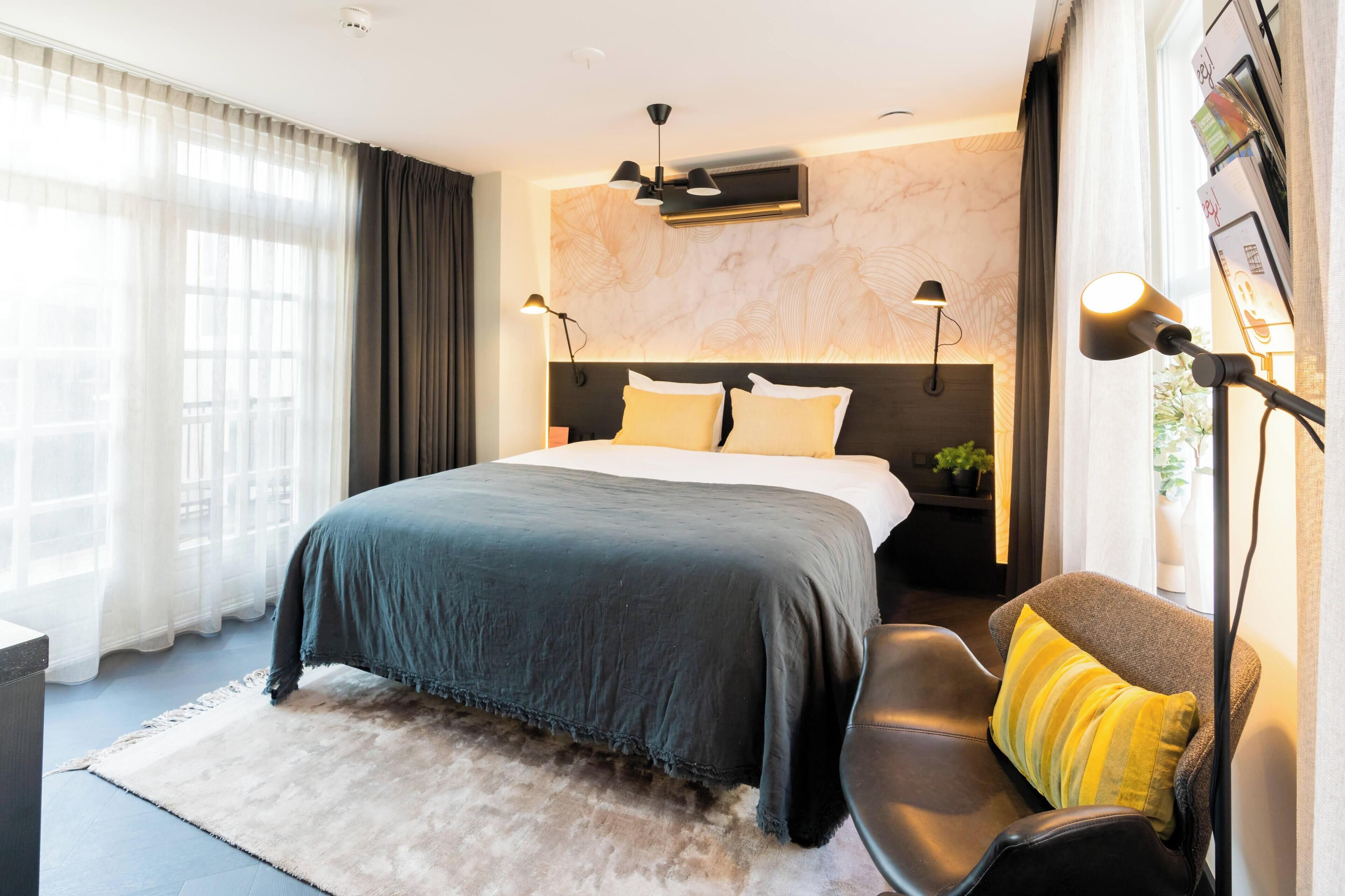 Quarto de hotel moderno com uma cama grande, uma escrivaninha e uma cadeira. Há duas mesas de cabeceira com luminárias, uma janela ampla com cortinas e um ar condicionado fixado na parede.
