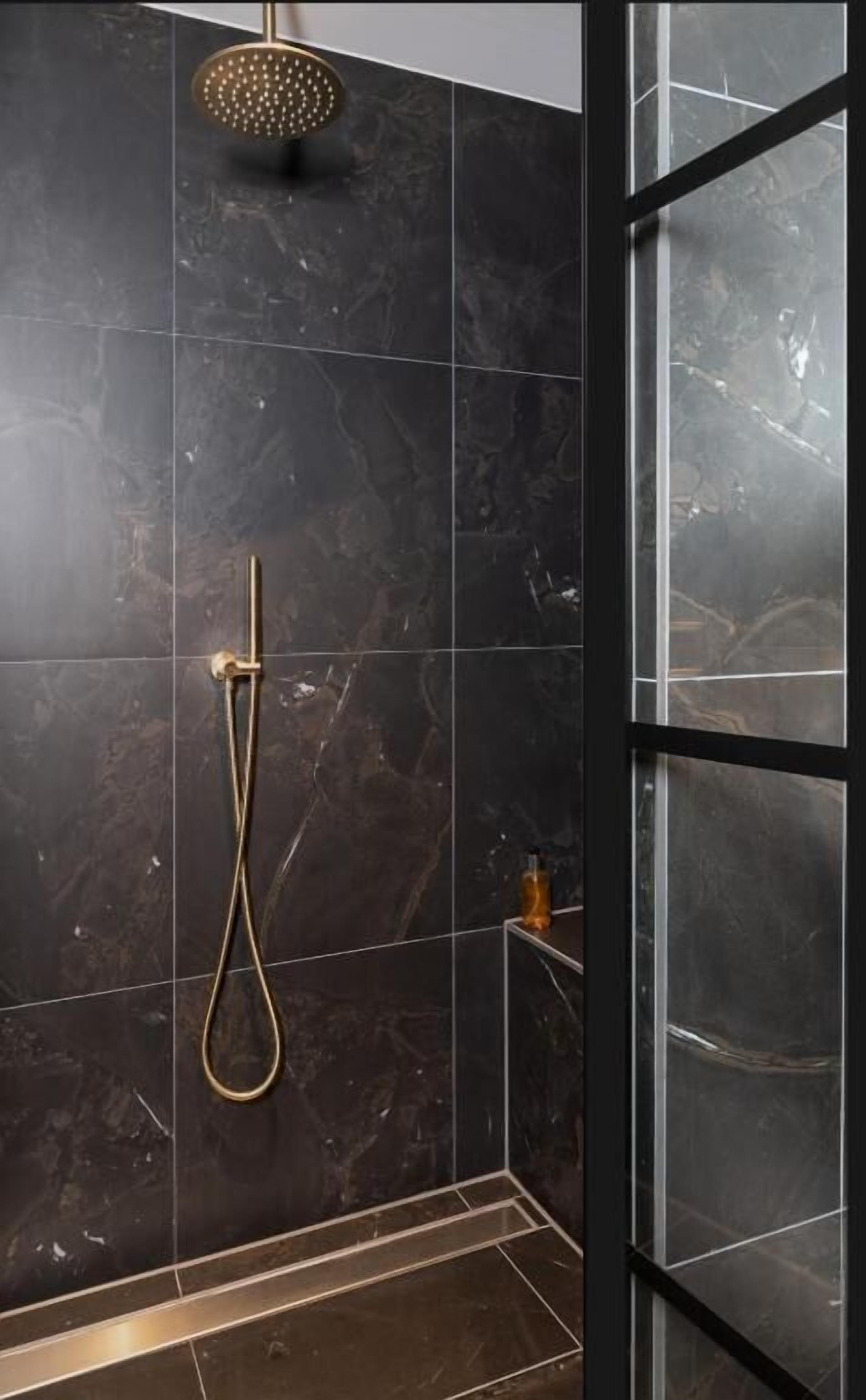Ducha moderna con cabezal de ducha dorado y cabezal de ducha de mano, sobre baldosas de mármol oscuro.