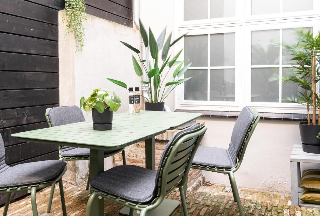 Ein Patio mit einem grünen Tisch und vier Stühlen, umgeben von Blumentöpfen und einem weißen Fenster.