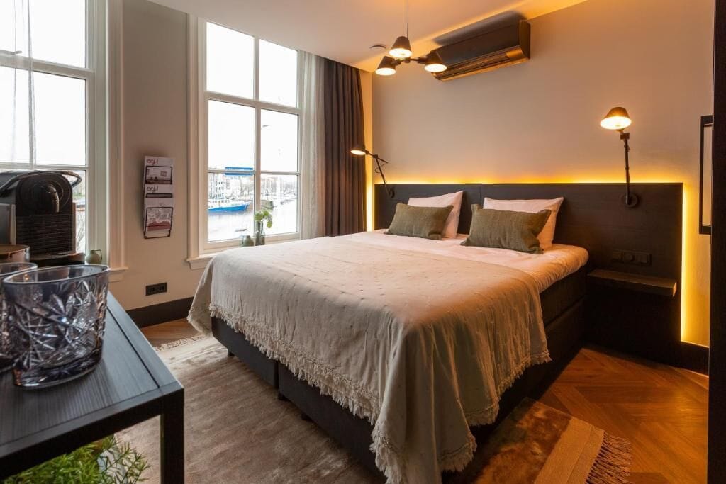 Une chambre d’hôtel moderne, dotée d’un grand lit, d’un bureau avec une lampe et offrant une vue sur la ville grâce à de grandes fenêtres.