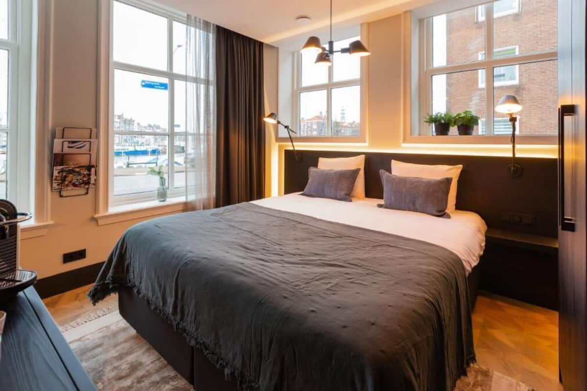 Ein modernes Hotelzimmer mit einem großen Bett, zwei Nachttischlampen, Blick auf die Stadt durch große Fenster und einem kleinen Tisch mit einem Korb.