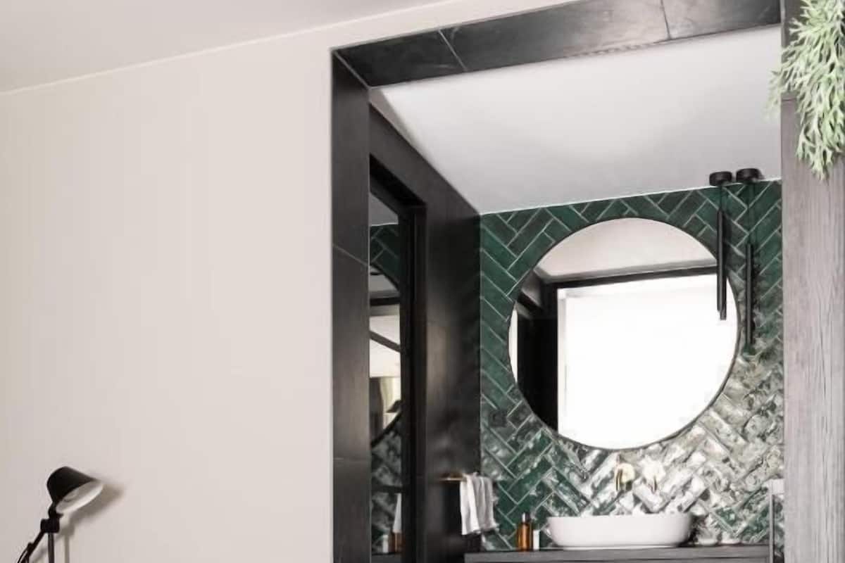 Ein modernes Schlafzimmer mit einem Bett, einer Nachttischlampe und Blick in ein Badezimmer mit großem Spiegel, dunklem Holzschrank und grün gemusterten Fliesen.