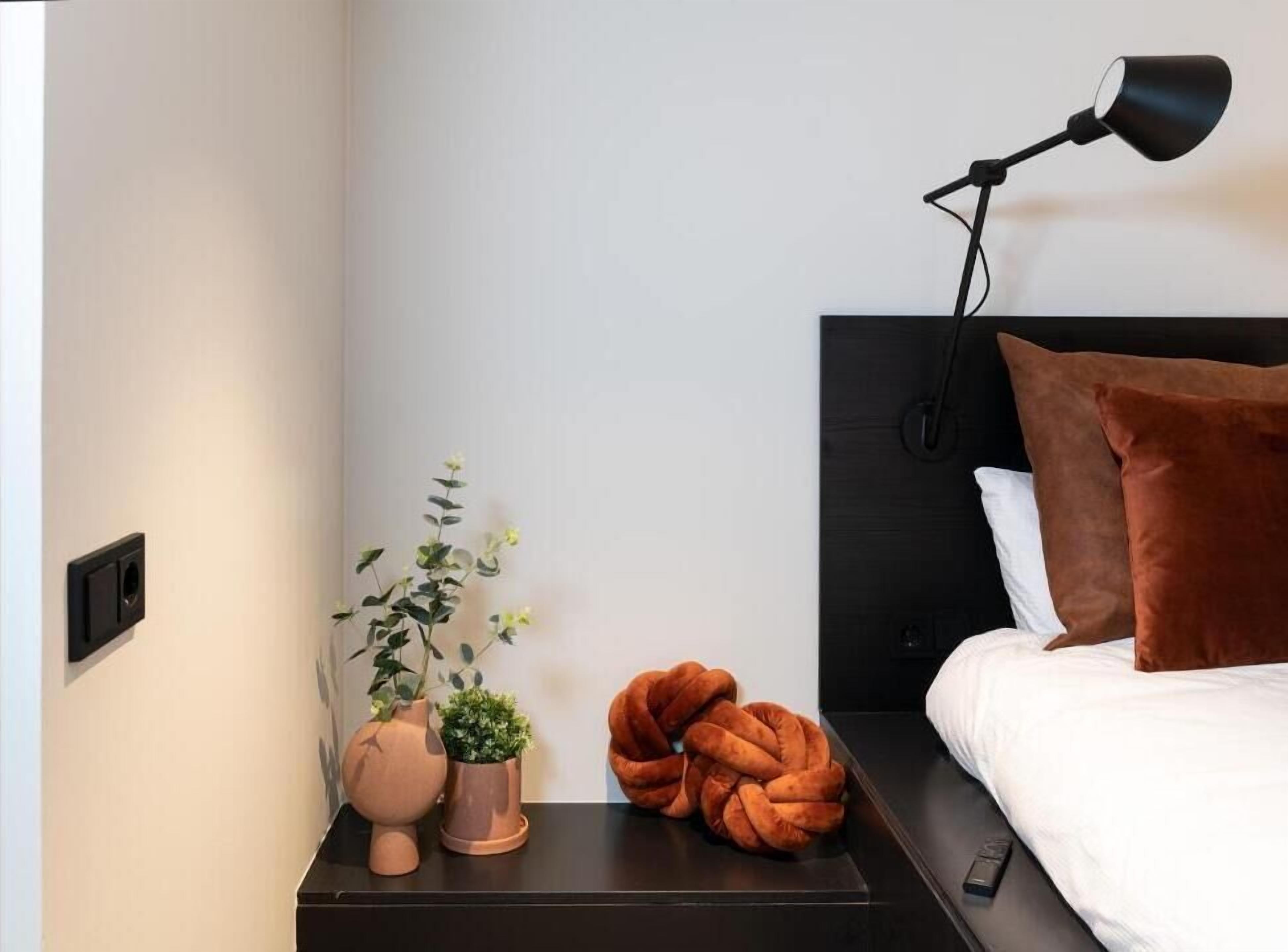 Ein ordentlich eingerichtetes Schlafzimmer mit einem schwarzen Kopfteil, einer Nachttischlampe, einem braunen Kissen, einem weißen Kissen, einem runden orangefarbenen Sitzkissen und zwei Topfpflanzen.
