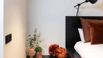 Ein ordentlich eingerichtetes Schlafzimmer mit einem schwarzen Kopfteil, einer Nachttischlampe, einem braunen Kissen, einem weißen Kissen, einem runden orangefarbenen Sitzkissen und zwei Topfpflanzen.