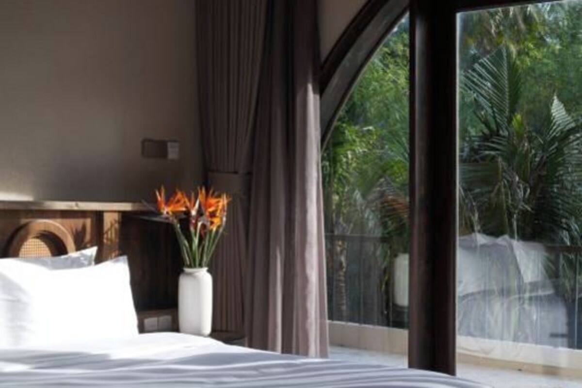 A hotel room with a bed, a vase with flowers, and a view of greenery through a large window.