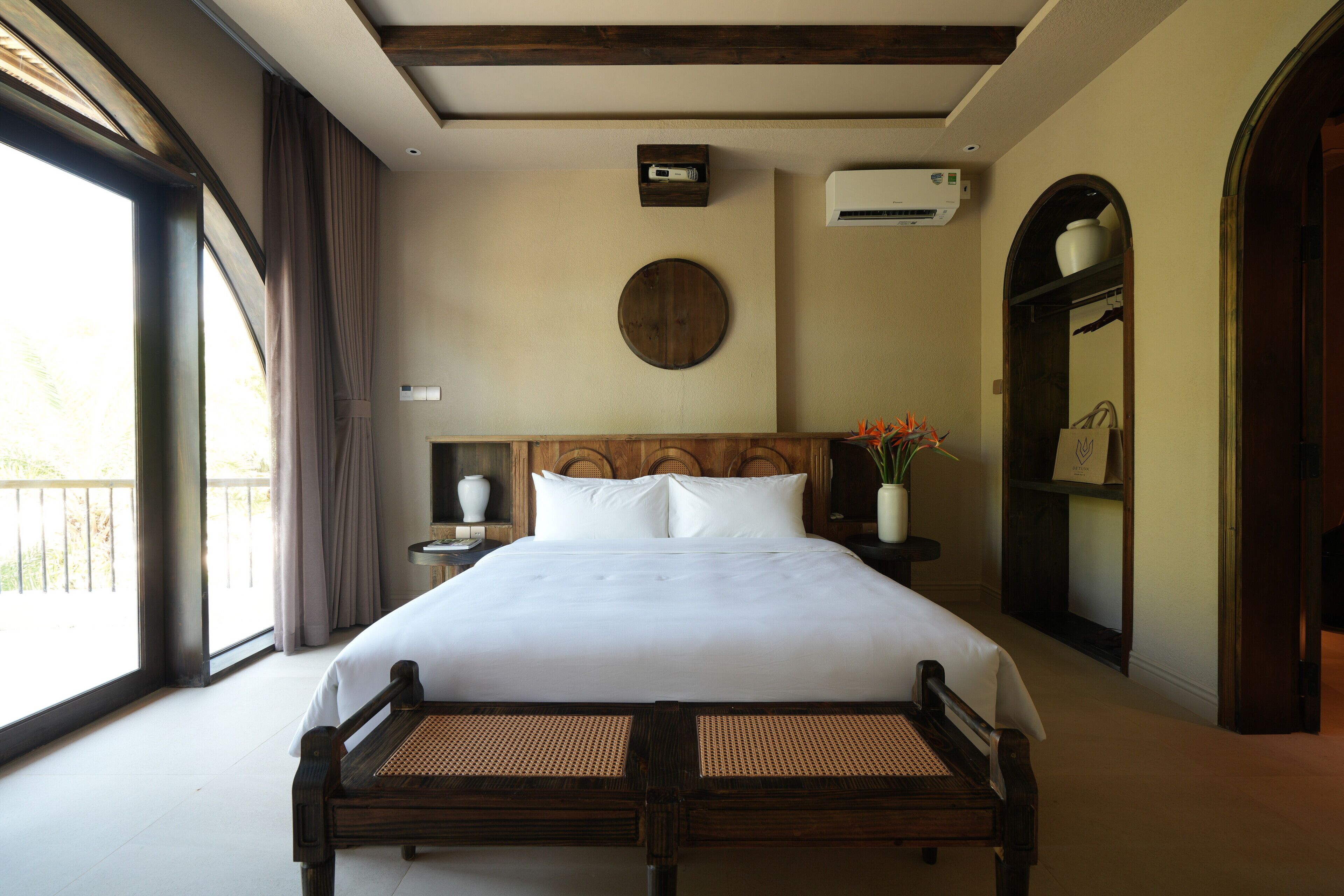 A neatly made bed with a wooden headboard, a bedside table with a lamp, and a large window with curtains.