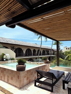 Un espace extérieur abrité comprenant un jacuzzi, des sièges et une vue sur un bâtiment doté d’arcades et d’un toit de chaume.