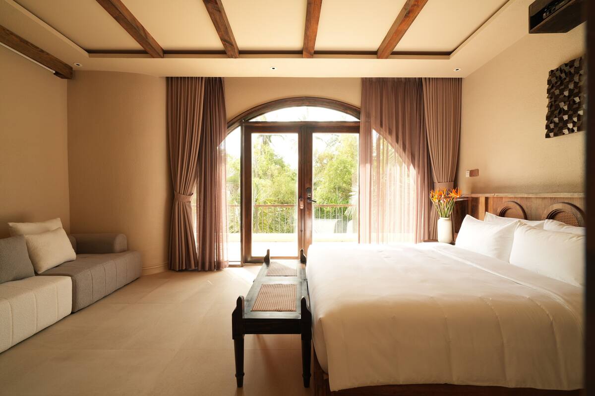 Une chambre d’hôtel avec un lit, un canapé, une petite table et une vue sur l’extérieur grâce à une grande fenêtre en arc de cercle.