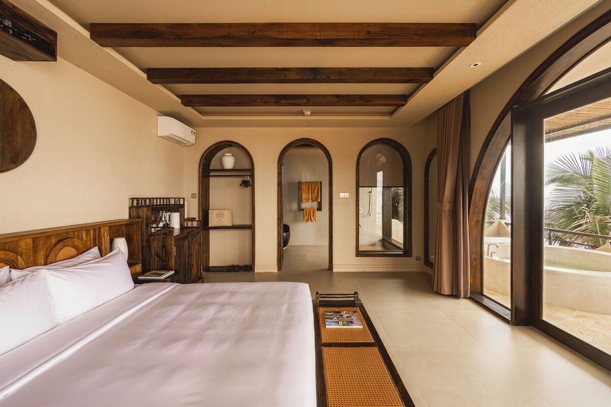 Une chambre d’hôtel avec un grand lit, des poutres apparentes en bois au plafond, une salle de bain avec baignoire et un balcon offrant une vue sur les palmiers.