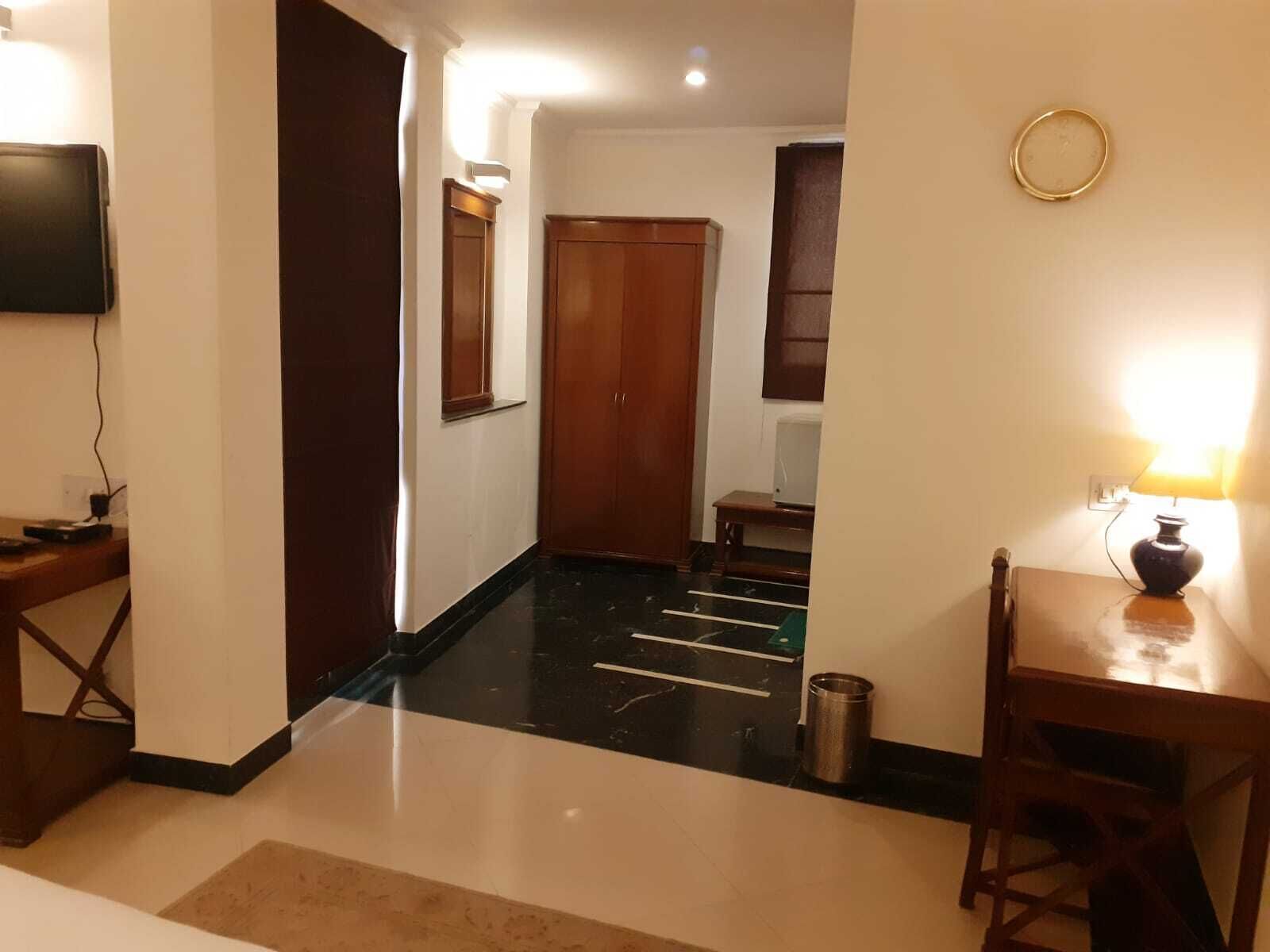 Una habitación de hotel con una puerta de madera, una mesita con una lámpara, un televisor montado en la pared y piso de baldosas.