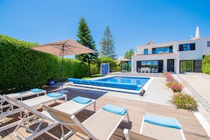 Un área moderna de piscina al aire libre con sillones de descanso y mesa de billar, rodeada por un seto cuidado y un edificio con grandes ventanales.