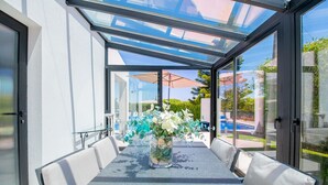 A glass-enclosed patio with a table and chairs, a vase with flowers, and a view of a pool and greenery. - 125 Quinta do Paraiso, elegant Holiday Villa! (Carvoeiro)