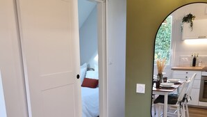 A room with a wooden floor, a white door, a bed with white and red bedding, and a kitchen area with a dining table and chairs.