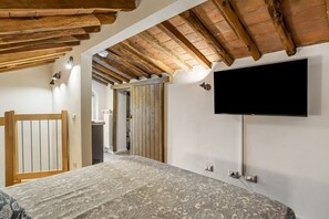 Quarto com cama, vigas de madeira no teto, televisão na parede e porta de acesso a outro cômodo.
