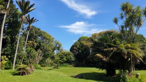 A lush garden with various tropical plants and trees, a well-maintained lawn, and a clear blue sky. - Huāki Punakaiki Bach (Punakaiki)