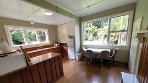 A kitchen with wooden cabinets, a built-in island, a dining area with chairs, and large windows with a view of greenery. - Huāki Punakaiki Bach (Punakaiki)