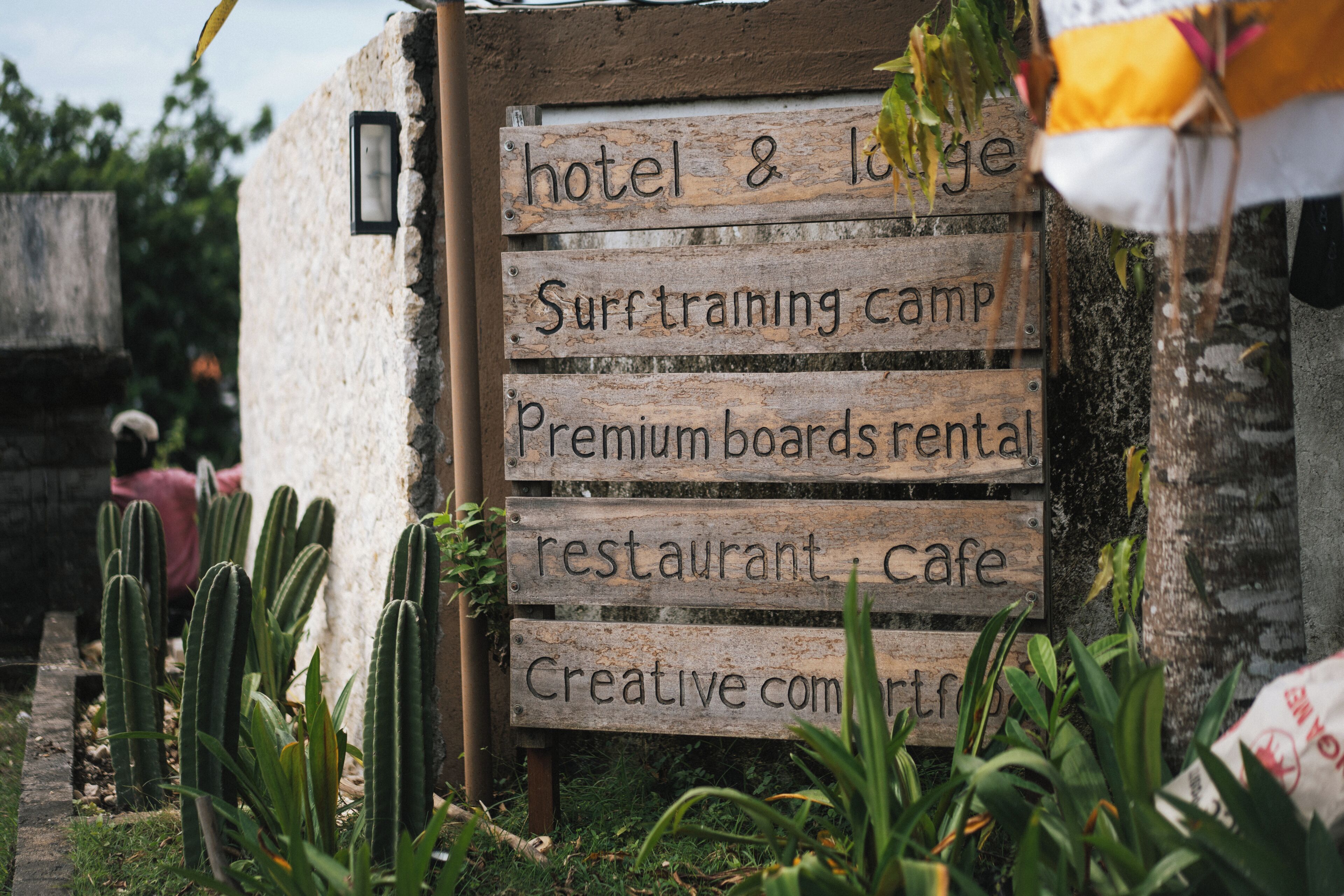Placa de madeira listando comodidades como hotel, acampamento de treino de surf, aluguer de pranchas premium, restaurante, café e comunidade criativa.