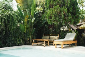 A poolside area with a wooden lounge chair and a small table, surrounded by lush greenery. - Olo Surf Nature (Jimbaran)