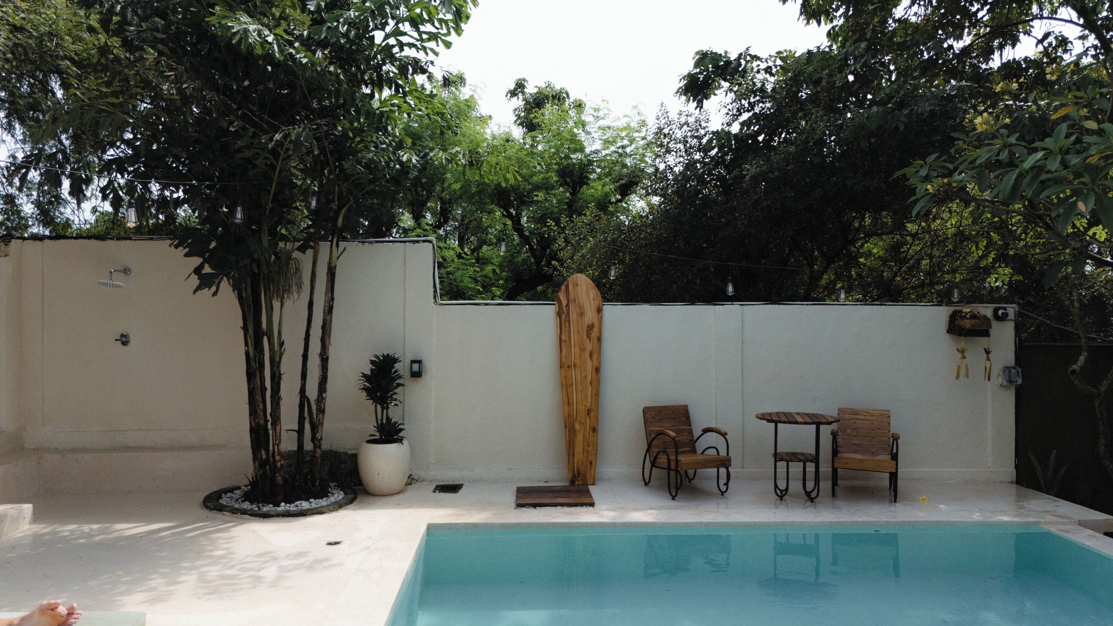 A swimming pool with two wooden chairs and a small table, surrounded by a white wall and trees.