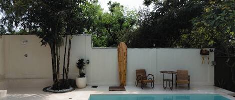 A swimming pool with two wooden chairs and a small table, surrounded by a white wall and trees.