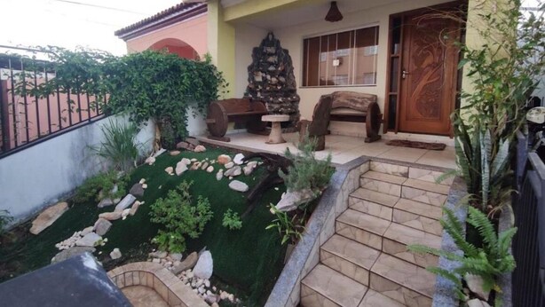 A house with a porch, a stone fireplace, and a small garden with rocks and plants. - Hostel Sideral (Campo Grande)