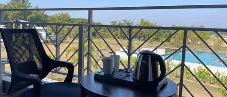 A balcony with two black chairs, a small table with a kettle and a mug, and a view of a swimming pool and greenery.