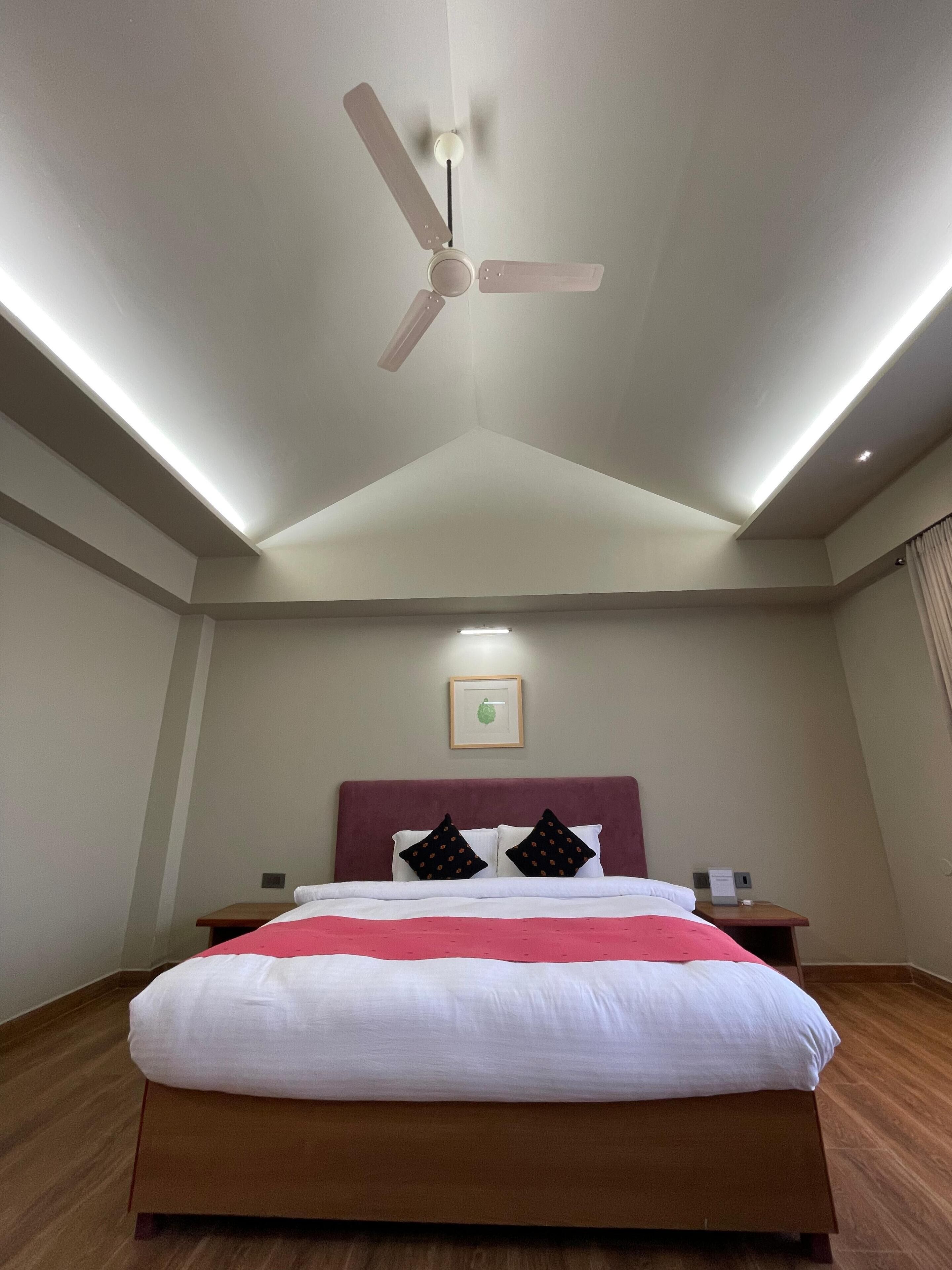 A hotel room with a bed, a ceiling fan, recessed lighting, a nightstand, a framed picture on the wall, and a wooden floor.