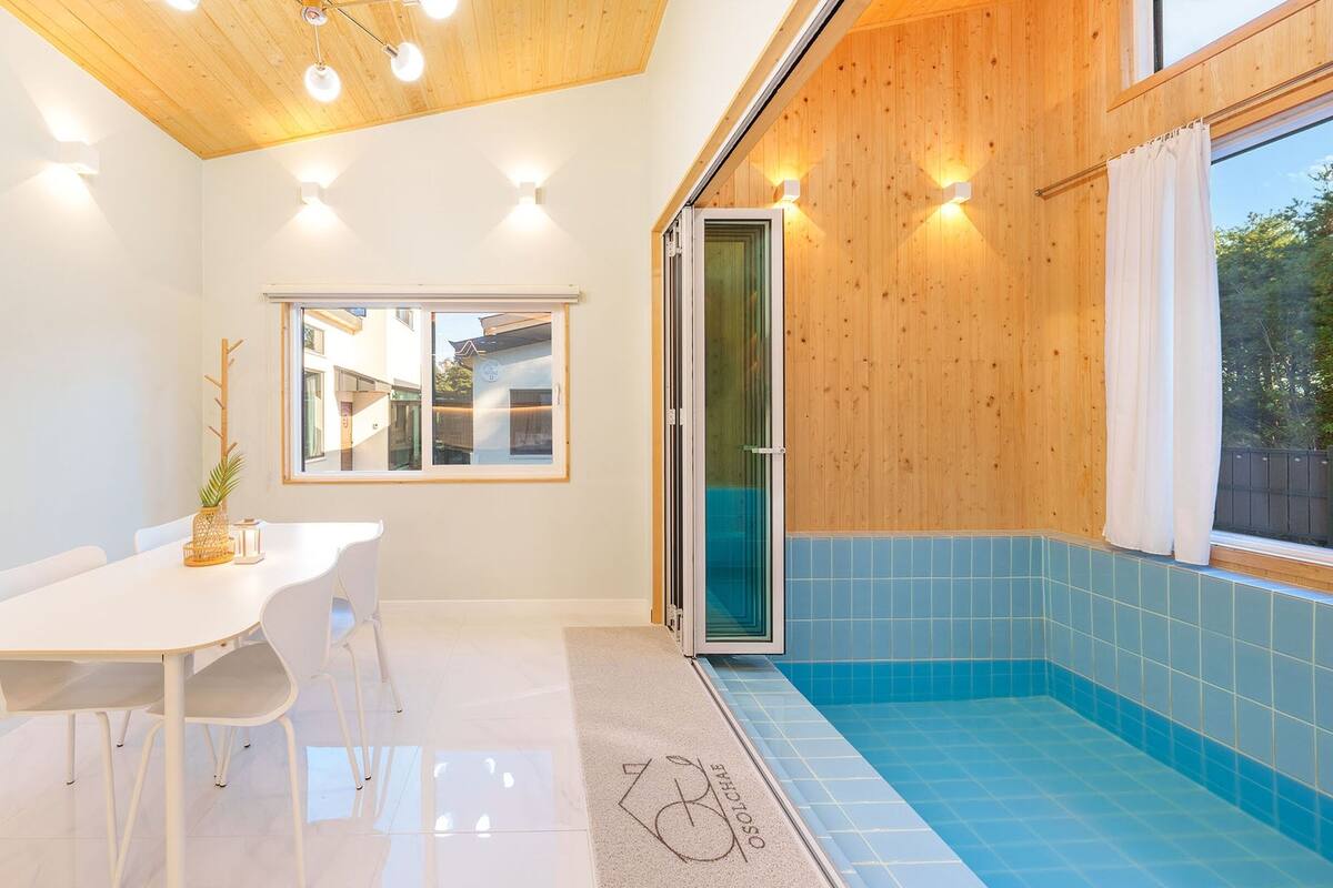 A modern kitchen with white cabinetry, a dining table with chairs, a window with a view of the outside, and a blue-tiled pool area.
