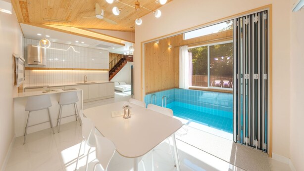 Uma cozinha moderna com armários brancos, uma área de refeições com mesa e cadeiras brancas, e uma grande porta de vidro deslizante que dá para uma área de piscina.
