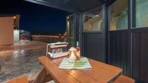 Um pátio externo com mesa de madeira posta para dois, uma garrafa de vinho e um candeeiro.