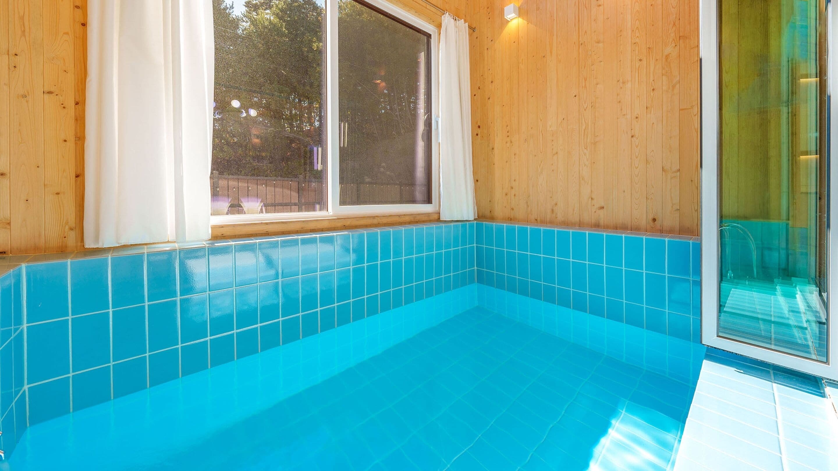 A small indoor swimming pool with blue tiles, wooden walls, and a window.