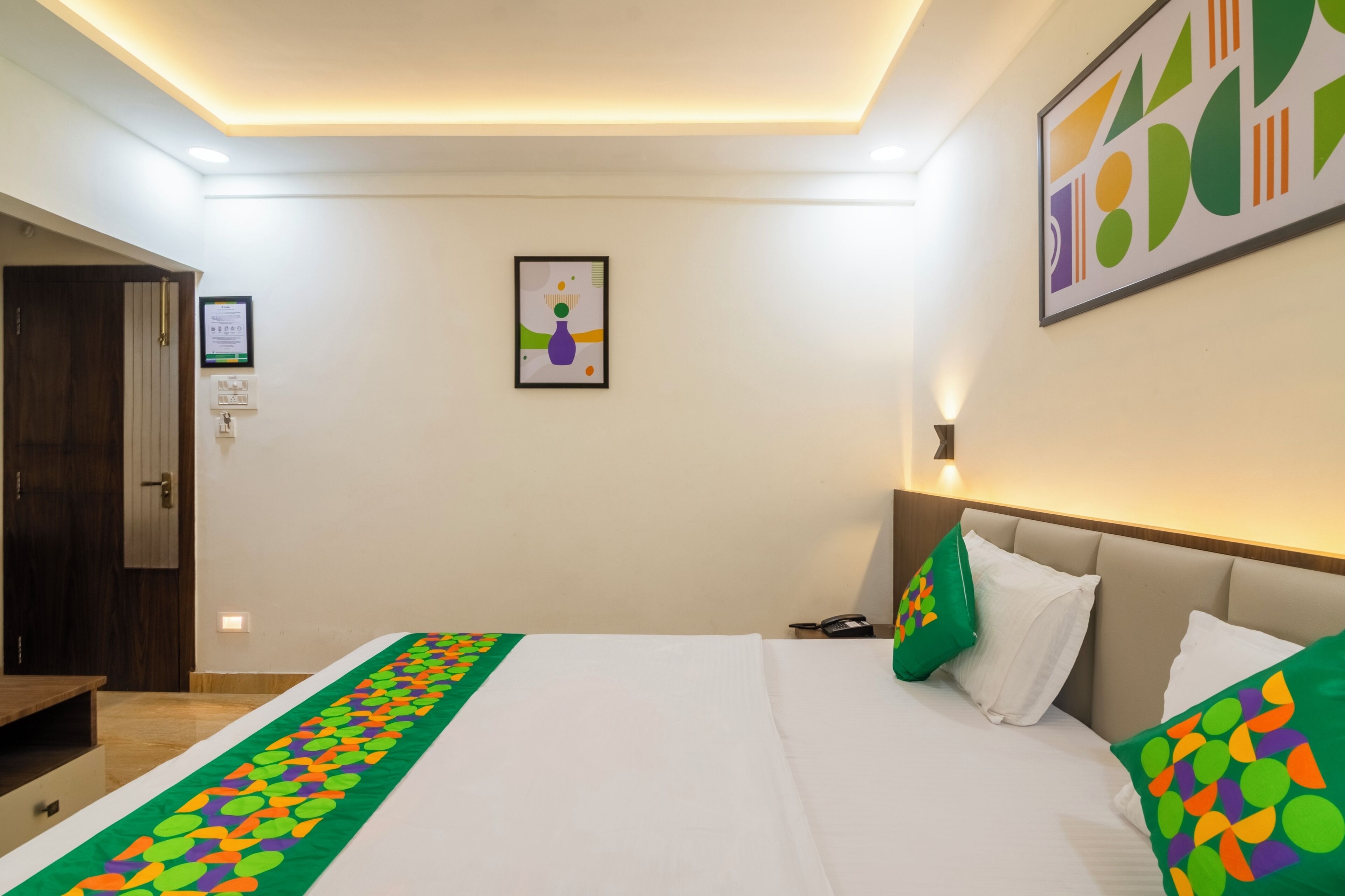 Une chambre d’hôtel avec un lit, une table de chevet, une commode, une lampe, un tableau encadré au mur et une porte.