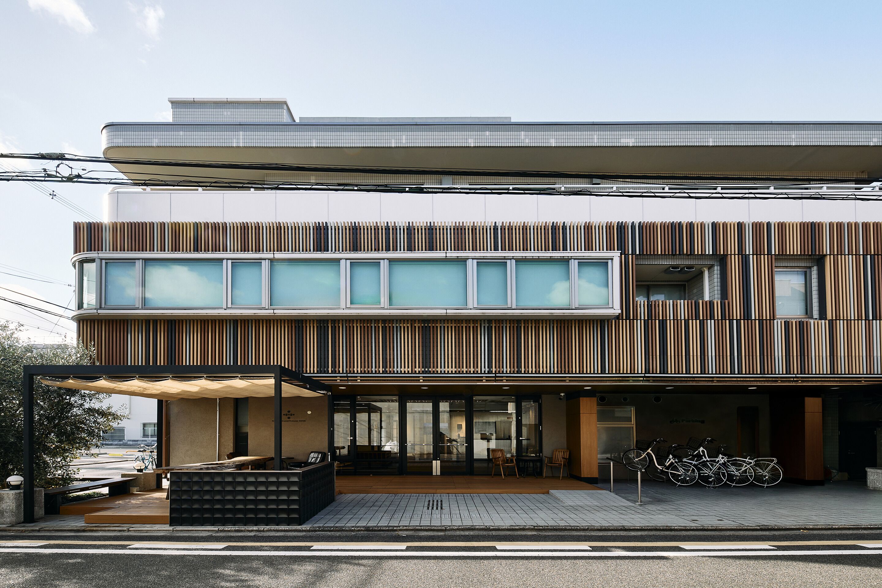 Un edificio moderno con techo plano, amplios ventanales y una zona de estar exterior cubierta donde se pueden ver bicicletas estacionadas cerca.
