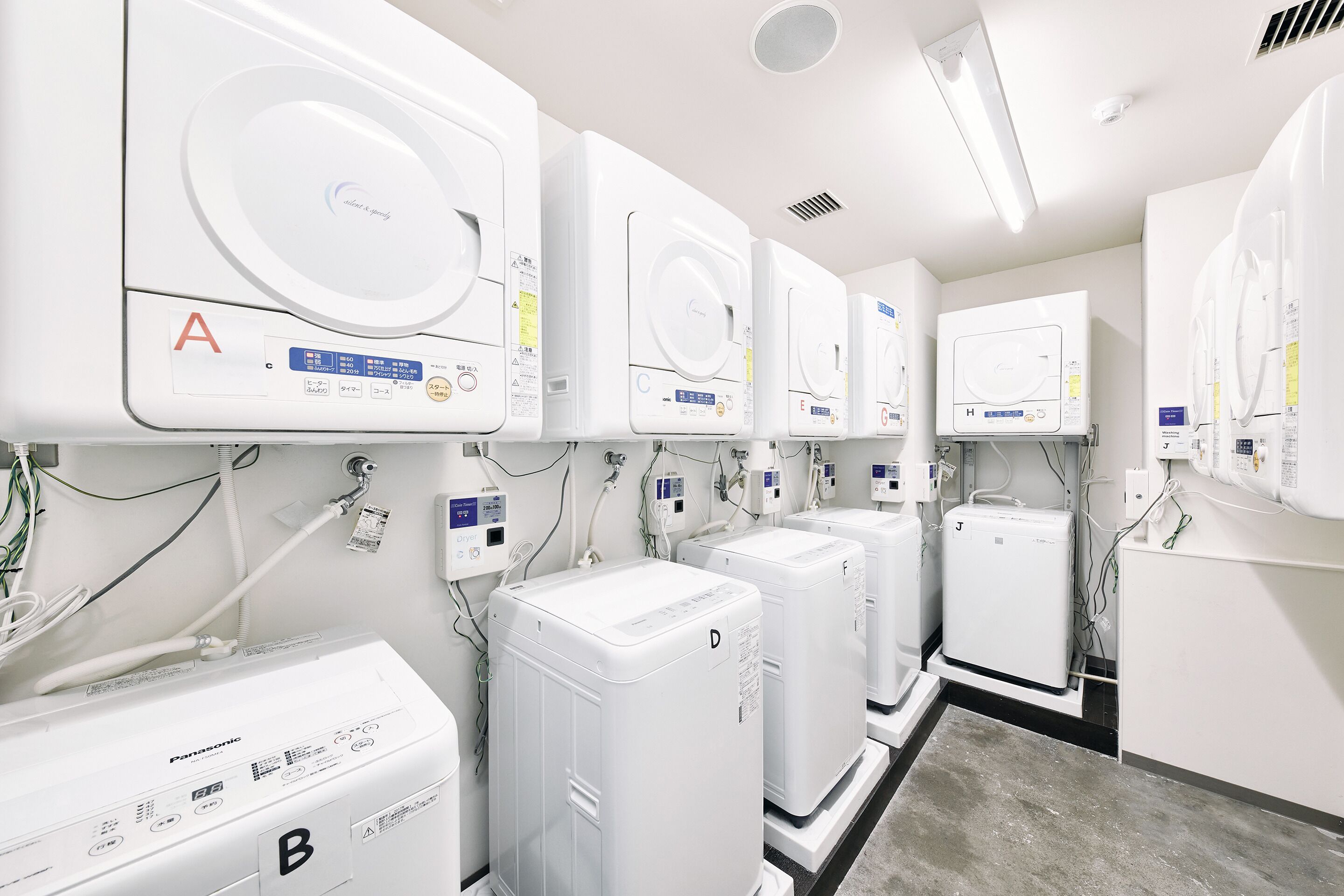 A laundry facility with multiple washing machines and dryers, labeled with letters A to H, and a control panel with various buttons and instructions.