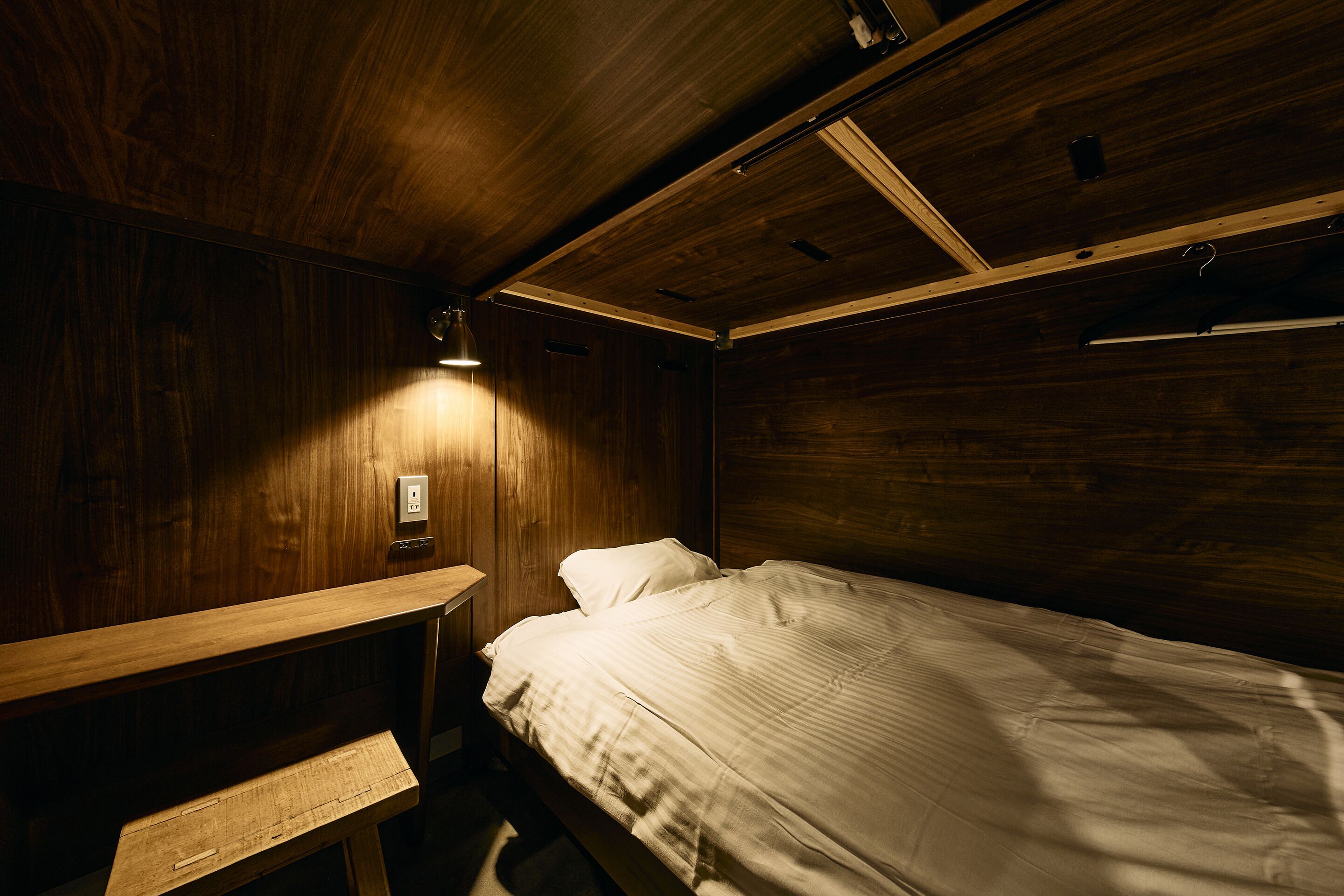 Una habitación de madera con una cama individual, un banco y una lámpara fijada a la pared.