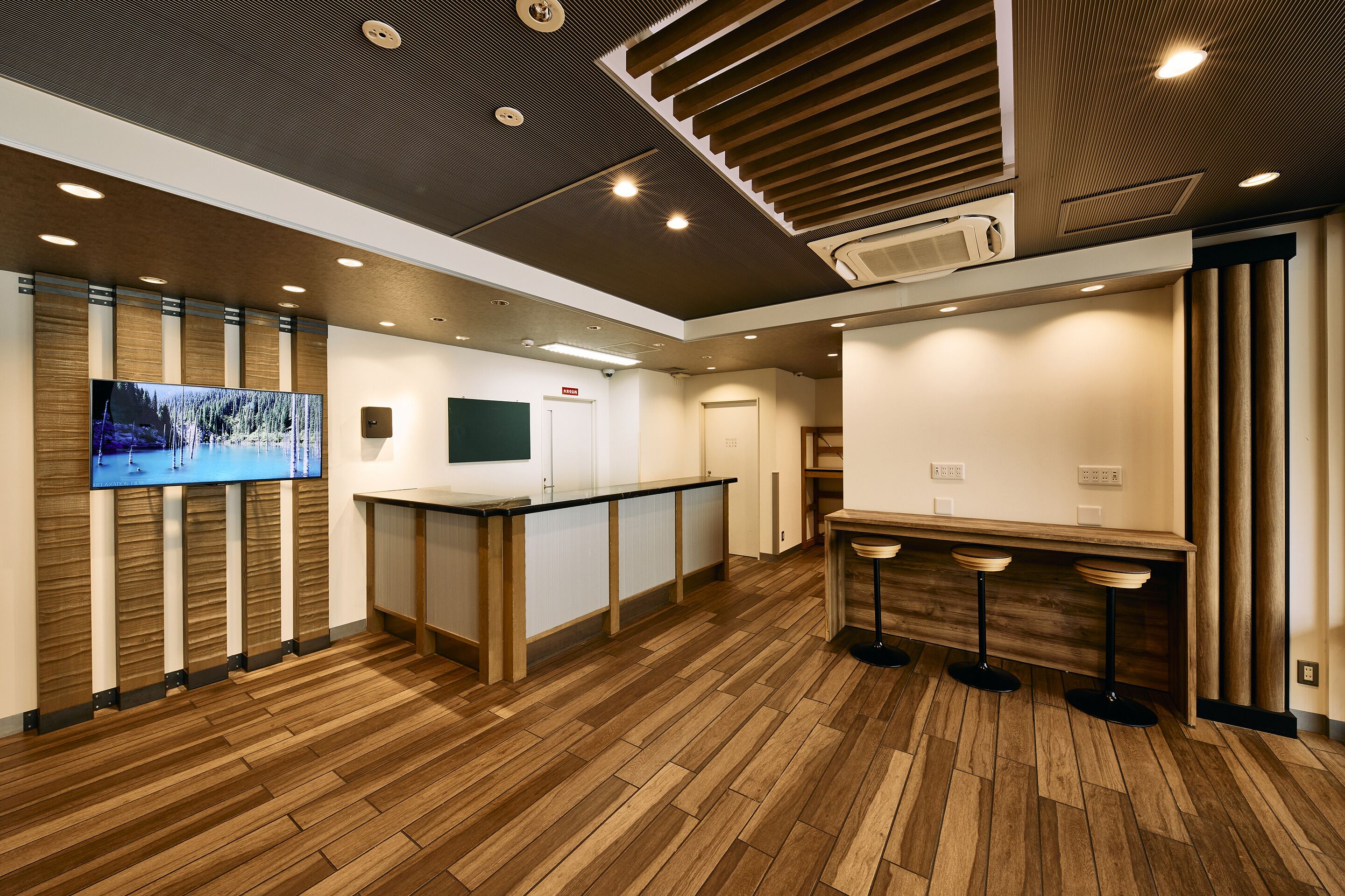 A modern reception area with a wooden floor, a reception desk, a flat-screen TV, and a bar area with stools.