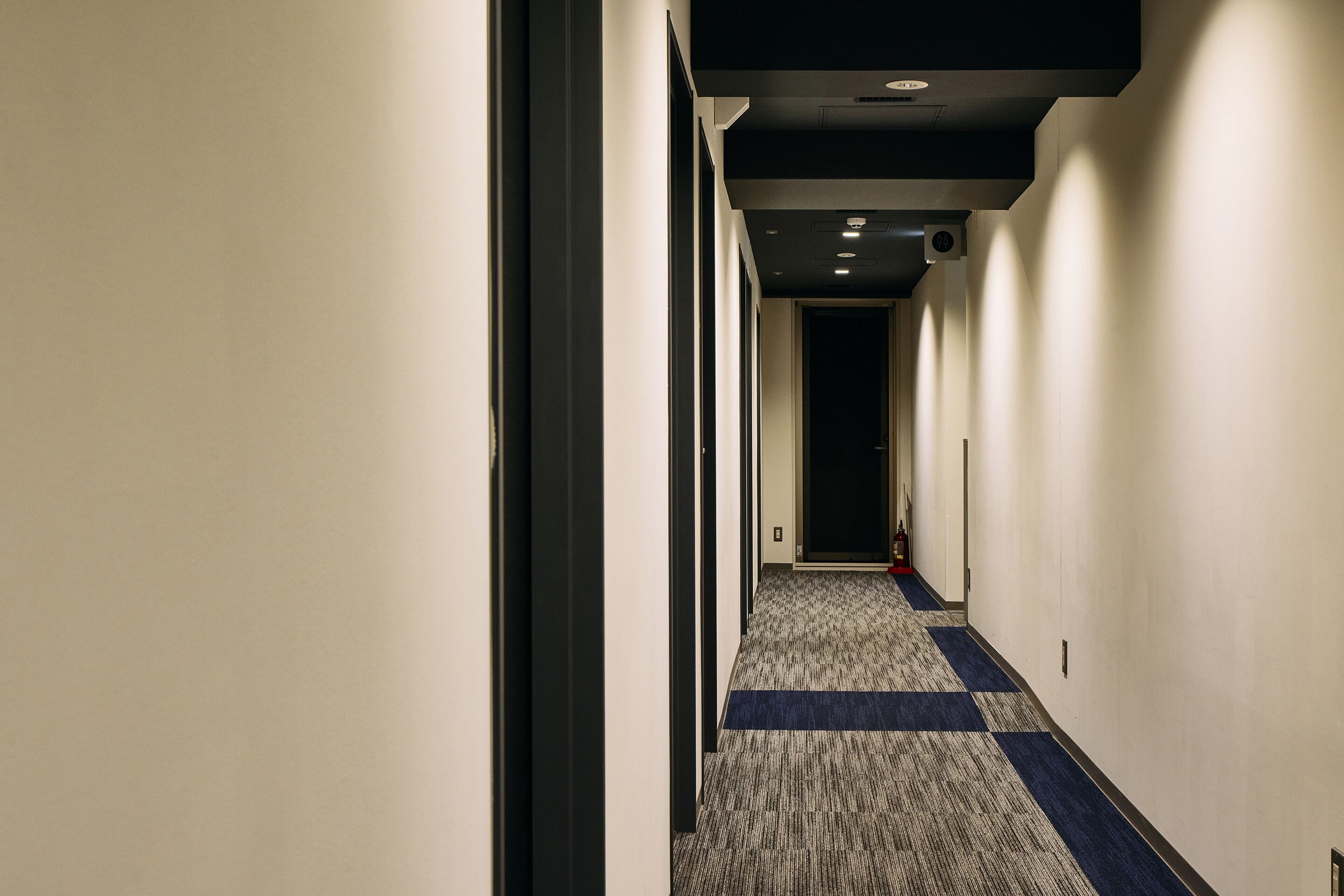 A long, narrow hotel corridor with a carpeted floor and a dark blue stripe running down the center.