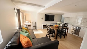 Una sala de estar moderna con un sofá negro, cojines coloridos, un comedor con mesa de madera y una cocina con armarios blancos y un lavavajillas de acero inoxidable.