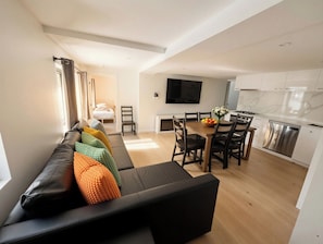 Una sala de estar moderna con un sofá negro, cojines coloridos, un comedor con mesa de madera y una cocina con armarios blancos y un lavavajillas de acero inoxidable.