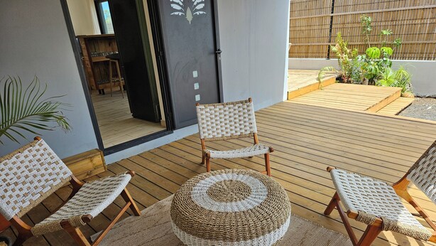 A wooden deck with wicker furniture, including chairs and a round table, and a view of the interior through a glass door. - Villa of the Three Monkeys. Between the beach and the mountains. (Saint-Pierre)