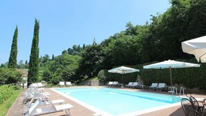 A rectangular swimming pool with a brick border, surrounded by lounge chairs and umbrellas, set against a backdrop of trees and a hill. - Nice apartment for 8 guests with pool, A/C, WIFI and TV, close to Verona (Colognola Ai Colli)